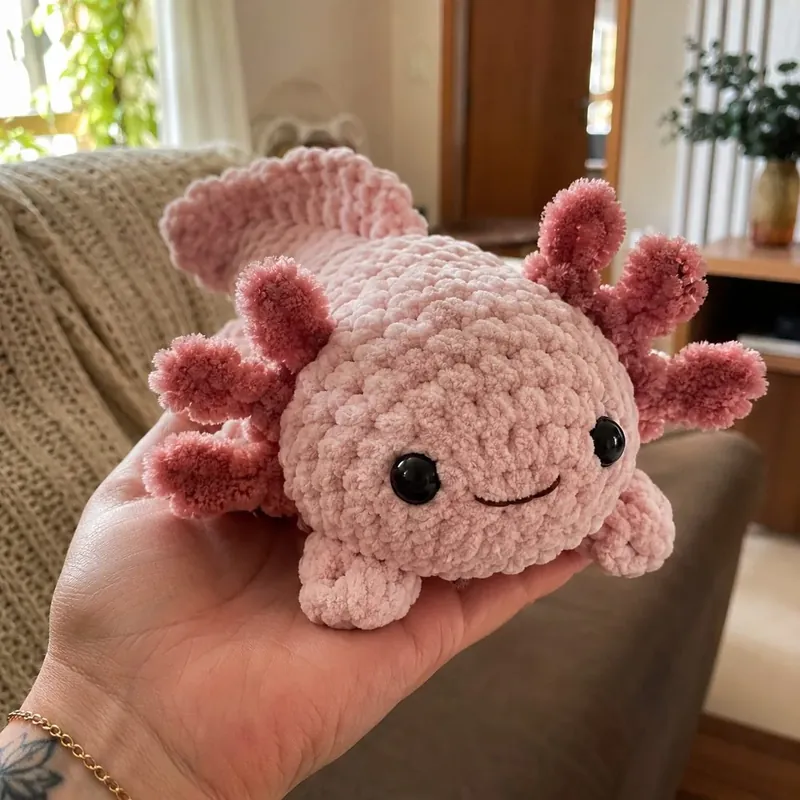 Ajolote rosa de crochet en hilo de terciopelo, apoyado en la palma de la mano.